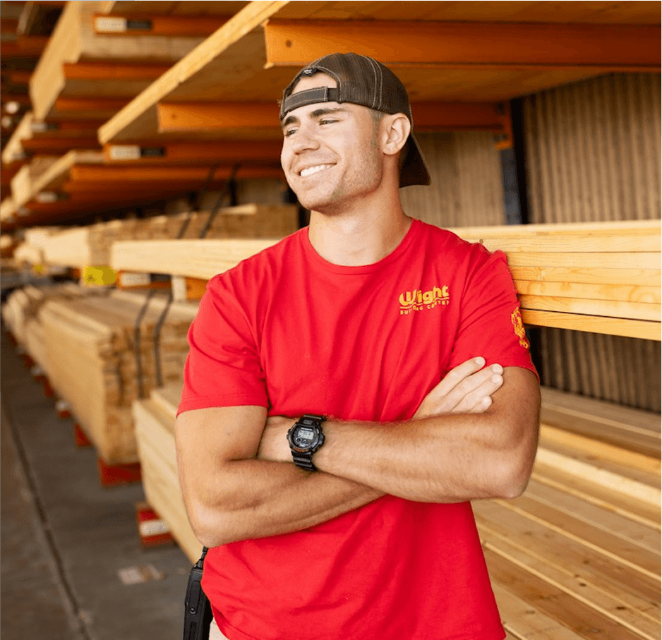 Smiling lumber yard worker in a red shirt, arms crossed, in front of stacked timber